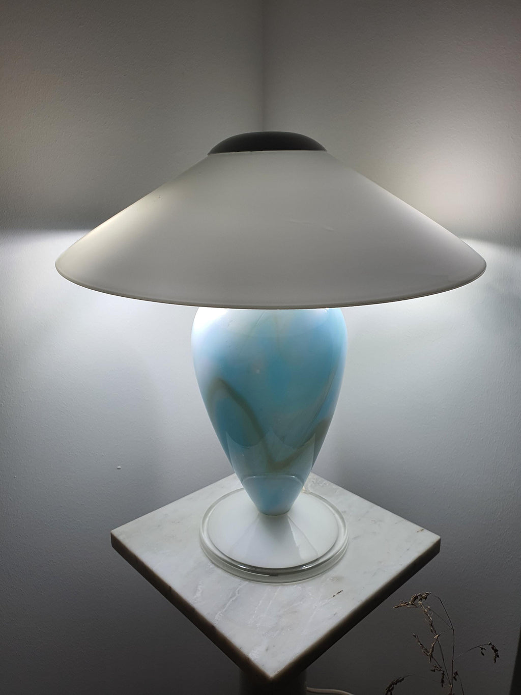 Front view of the lit Murano glass mushroom table lamp, sky blue marbled glass base beneath a wide white glass shade, on a marble pedestal.