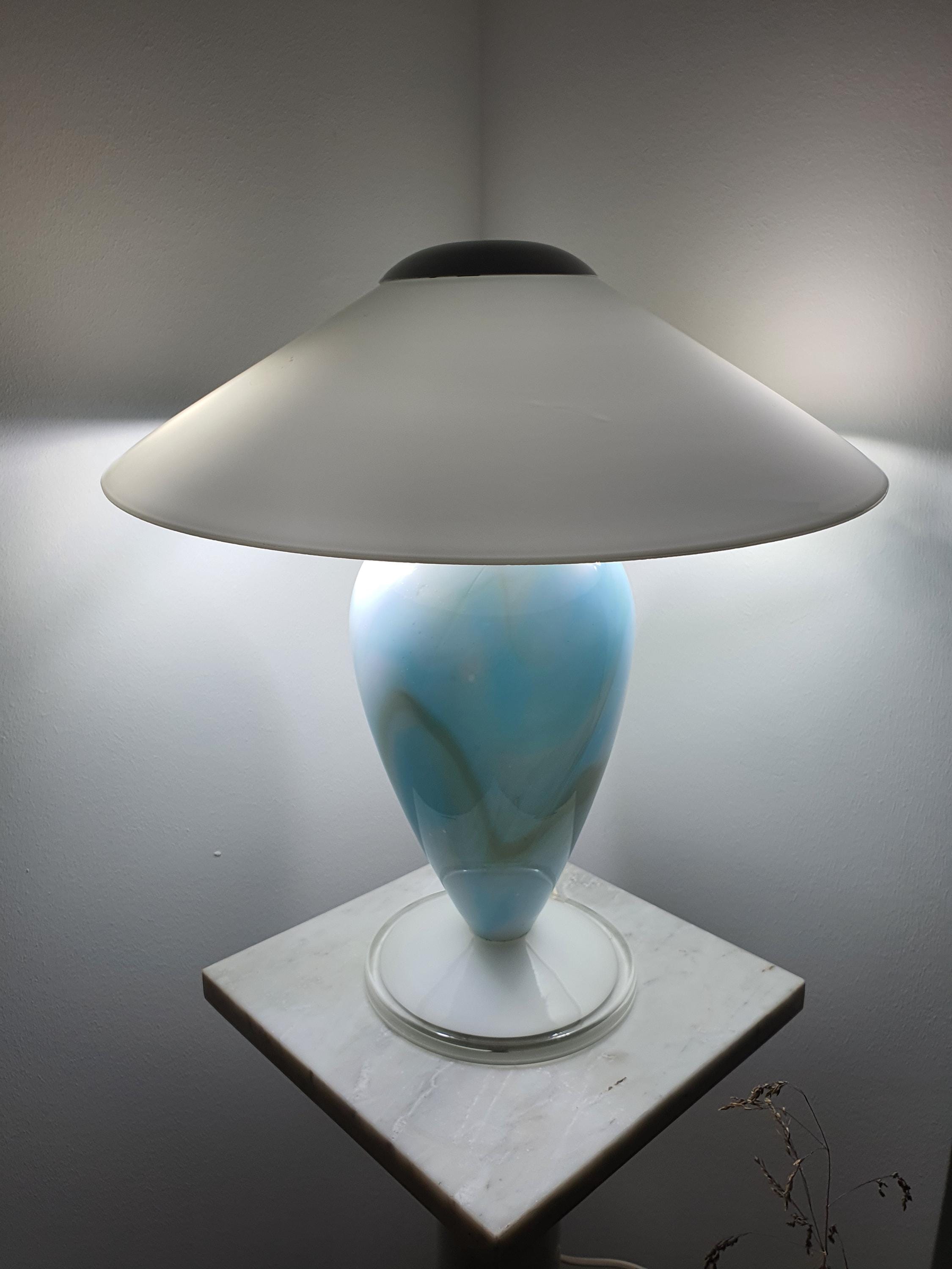 Front view of the lit Murano glass mushroom table lamp, sky blue marbled glass base beneath a wide white glass shade, on a marble pedestal.