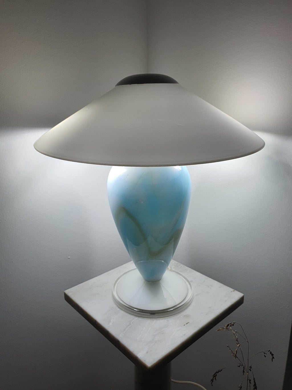 Full view of the illuminated sky blue marbled Murano glass mushroom table lamp, showing the wide white shade and glowing glass base on a marble stand.