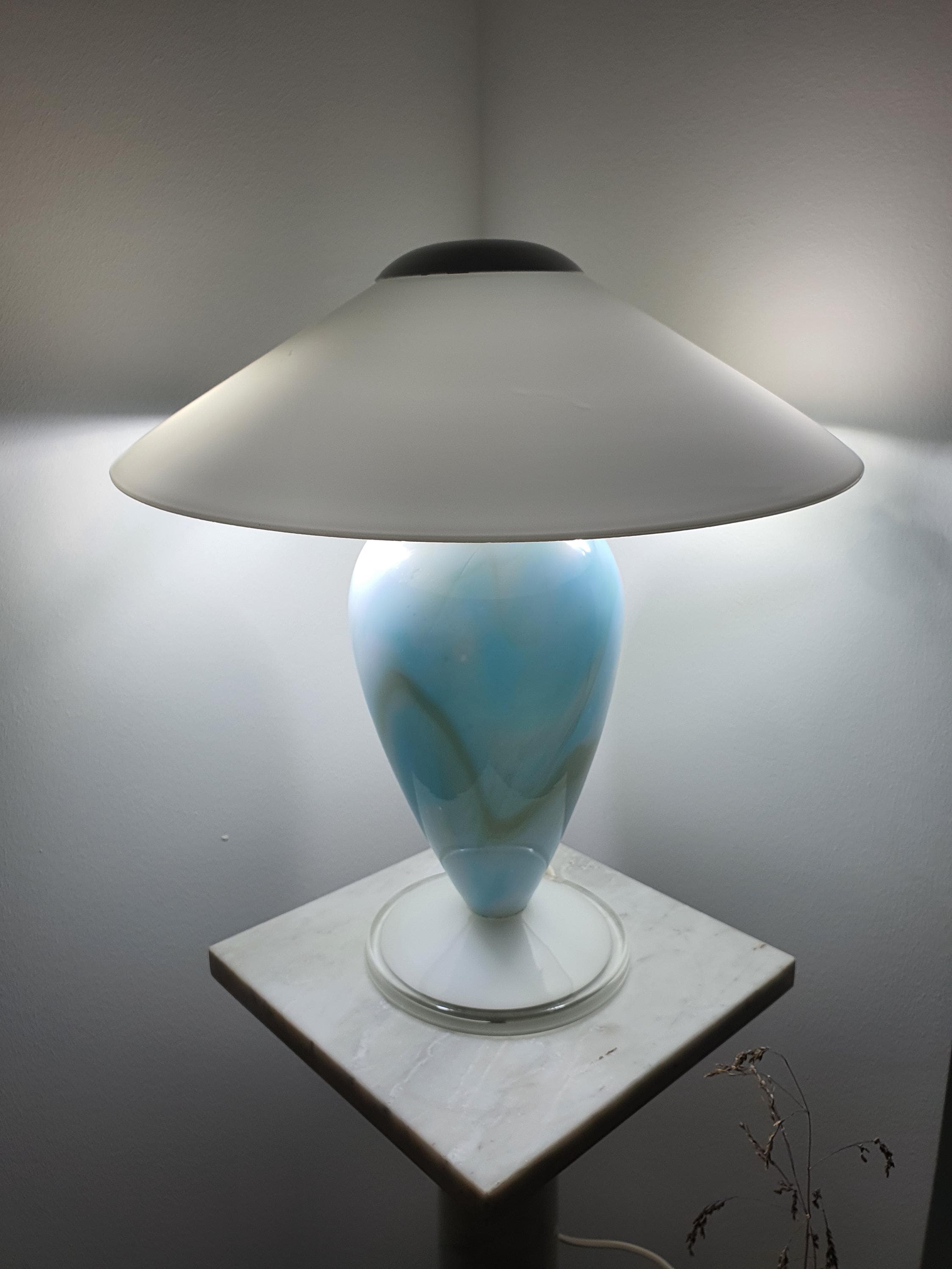 Full view of the illuminated sky blue marbled Murano glass mushroom table lamp, showing the wide white shade and glowing glass base on a marble stand.