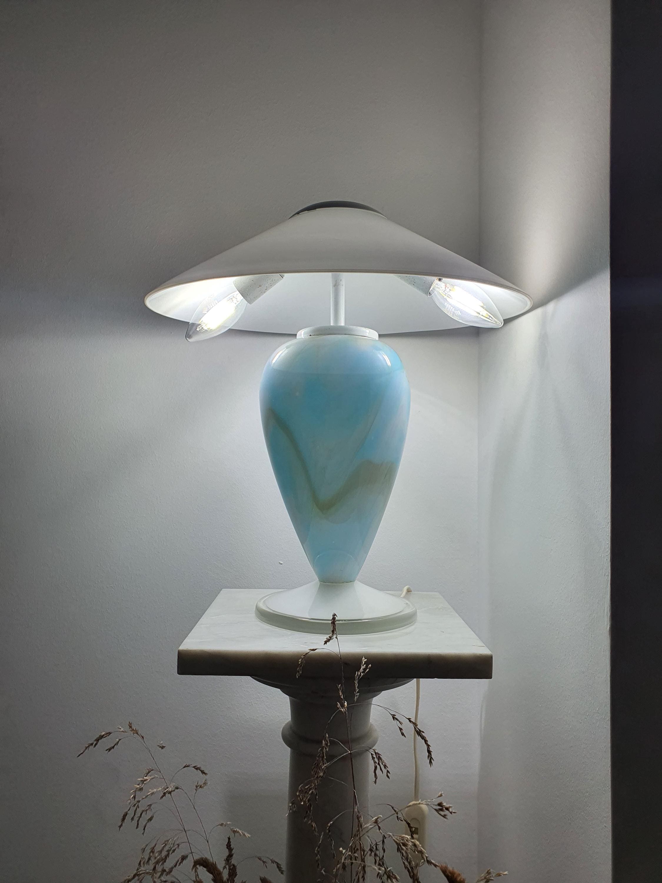 Sky blue marbled Murano glass mushroom table lamp lit and glowing, showing light diffused through the white glass shade, displayed on a marble pedestal.