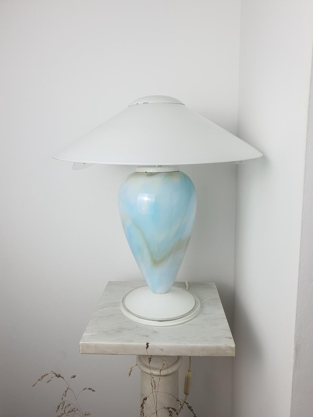 Sky blue marbled Murano glass mushroom table lamp with white conical shade, viewed from a side angle on a marble pedestal against a white wall.