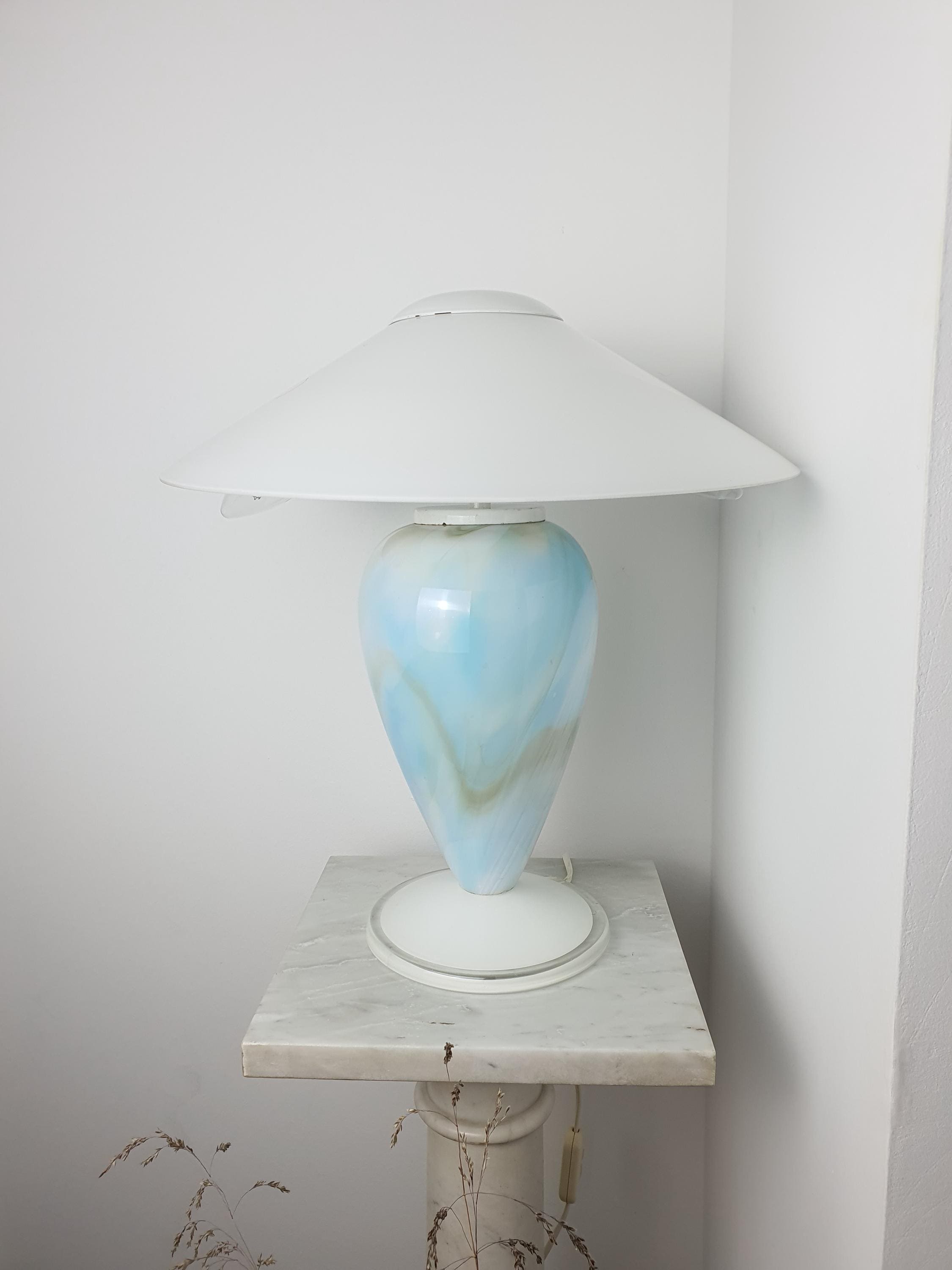 Sky blue marbled Murano glass mushroom table lamp with white conical shade, viewed from a side angle on a marble pedestal against a white wall.