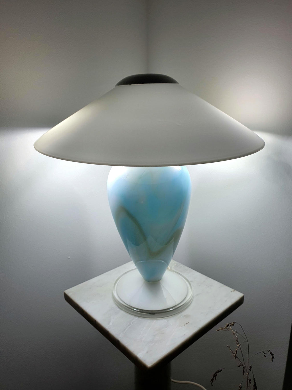 Sky blue marbled Murano glass mushroom table lamp with rounded white glass shade, lit and displayed on a marble pedestal against a grey wall.