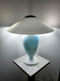 Sky blue marbled Murano glass mushroom table lamp with rounded white glass shade, lit and displayed on a marble pedestal against a grey wall.
