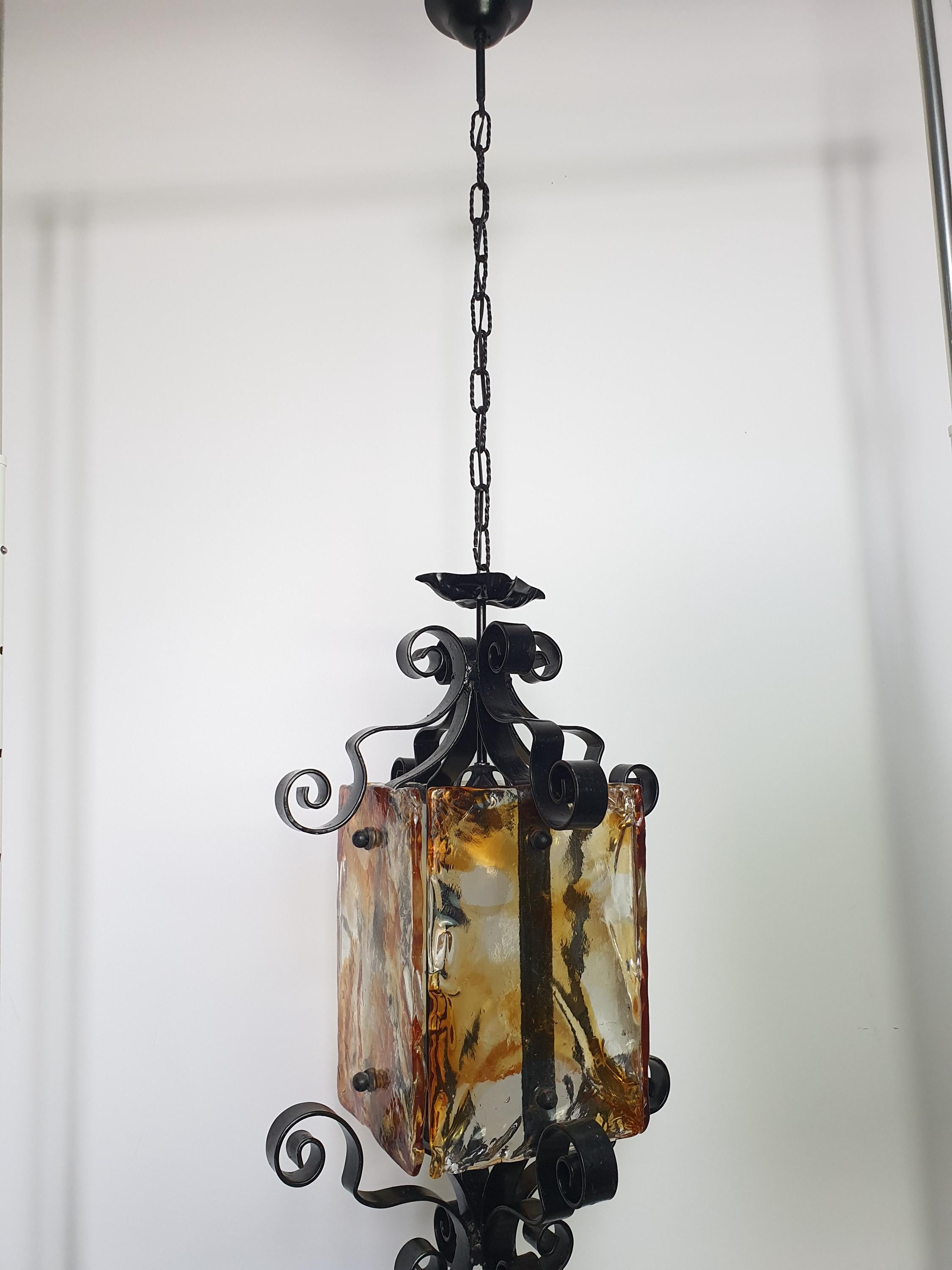 Mazzega style Murano pendant lit, warm golden amber glow, dramatic dark shadows, black wrought iron frame, high ceiling setting