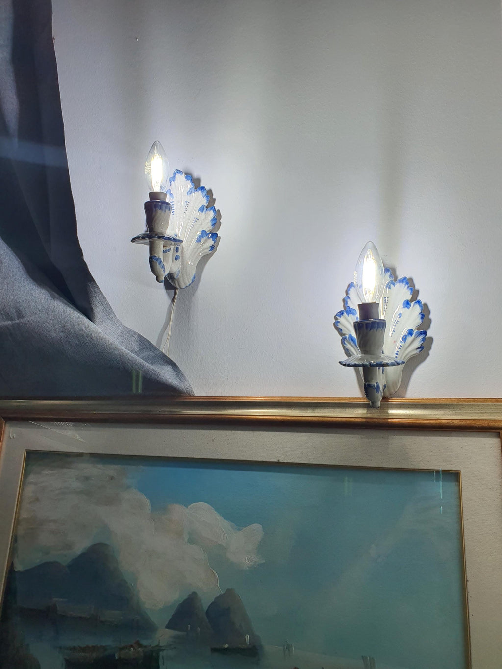 Pair of vintage Italian Bassano ceramic wall sconces lit, warm soft glow, white glaze and blue hand-painted details, cottage interior